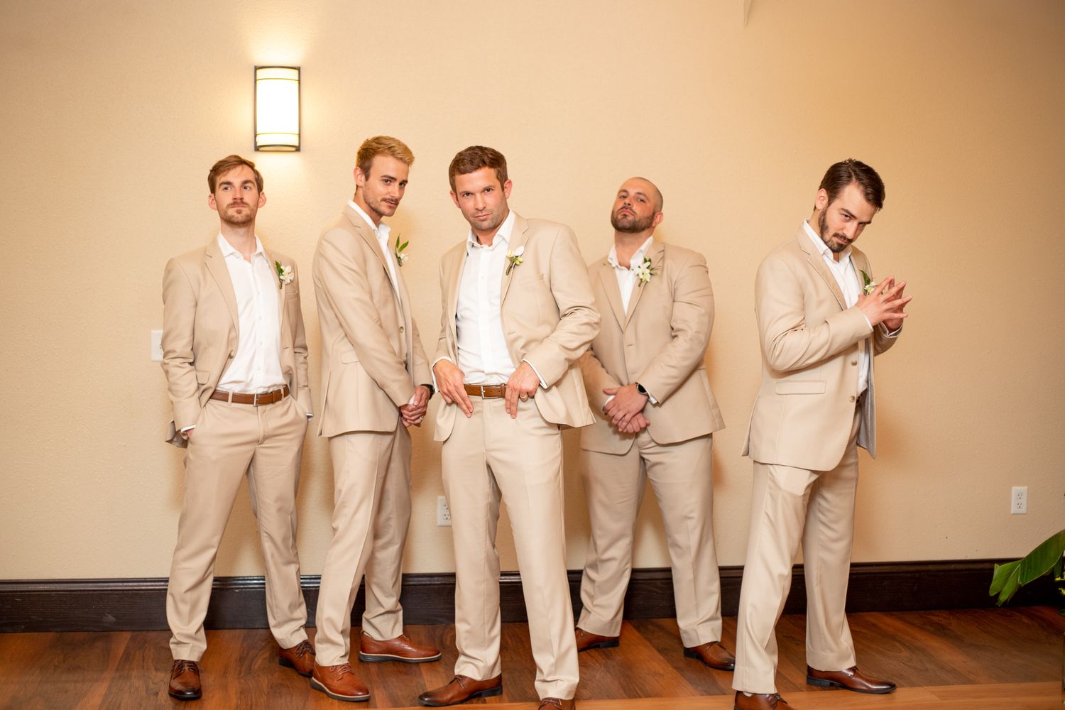 Five groomsmen in matching tan suits with boutonnieres posing indoors at a wedding venue.