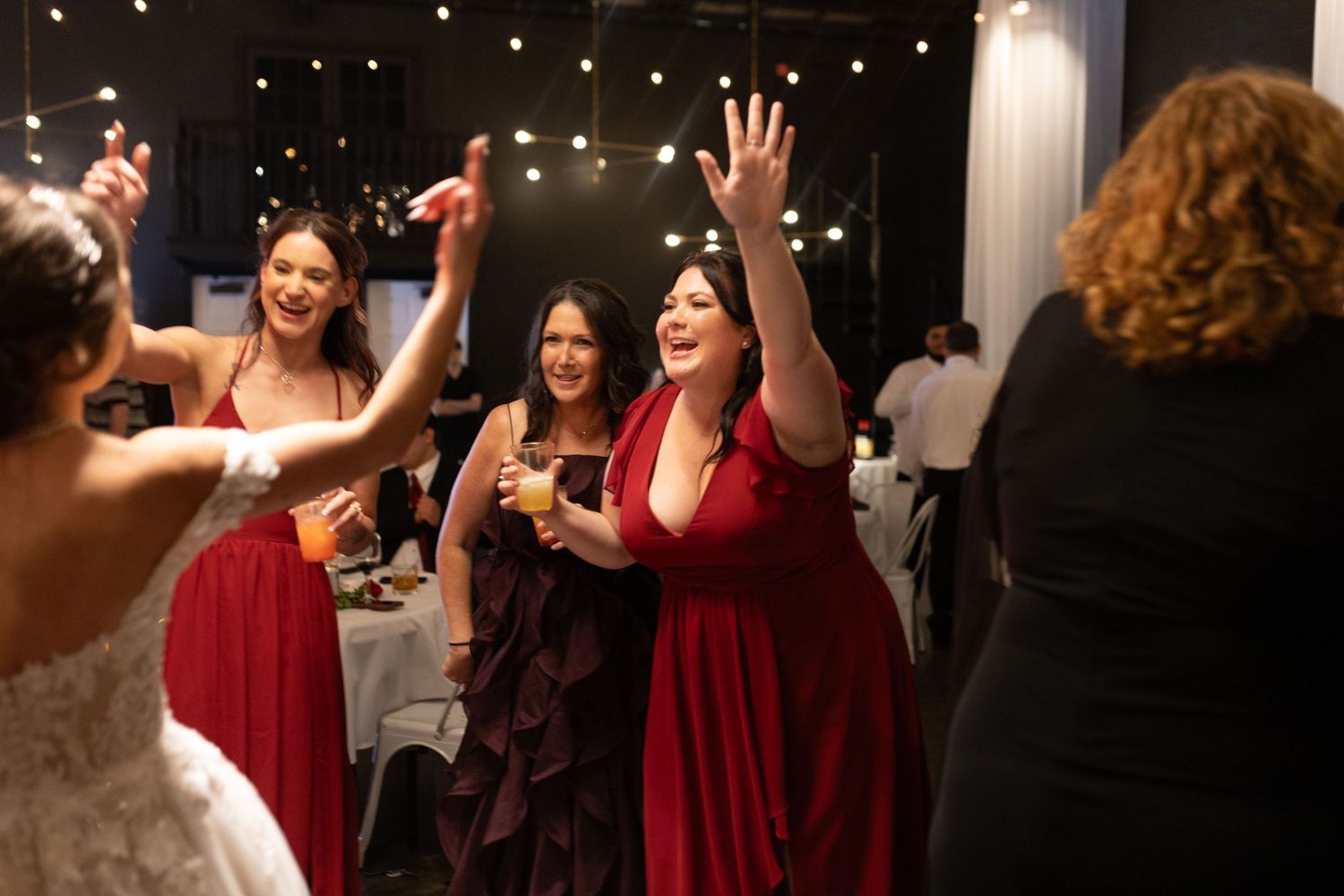 Women in red dresses dancing and celebrating joyfully at a wedding reception with string lights in background.