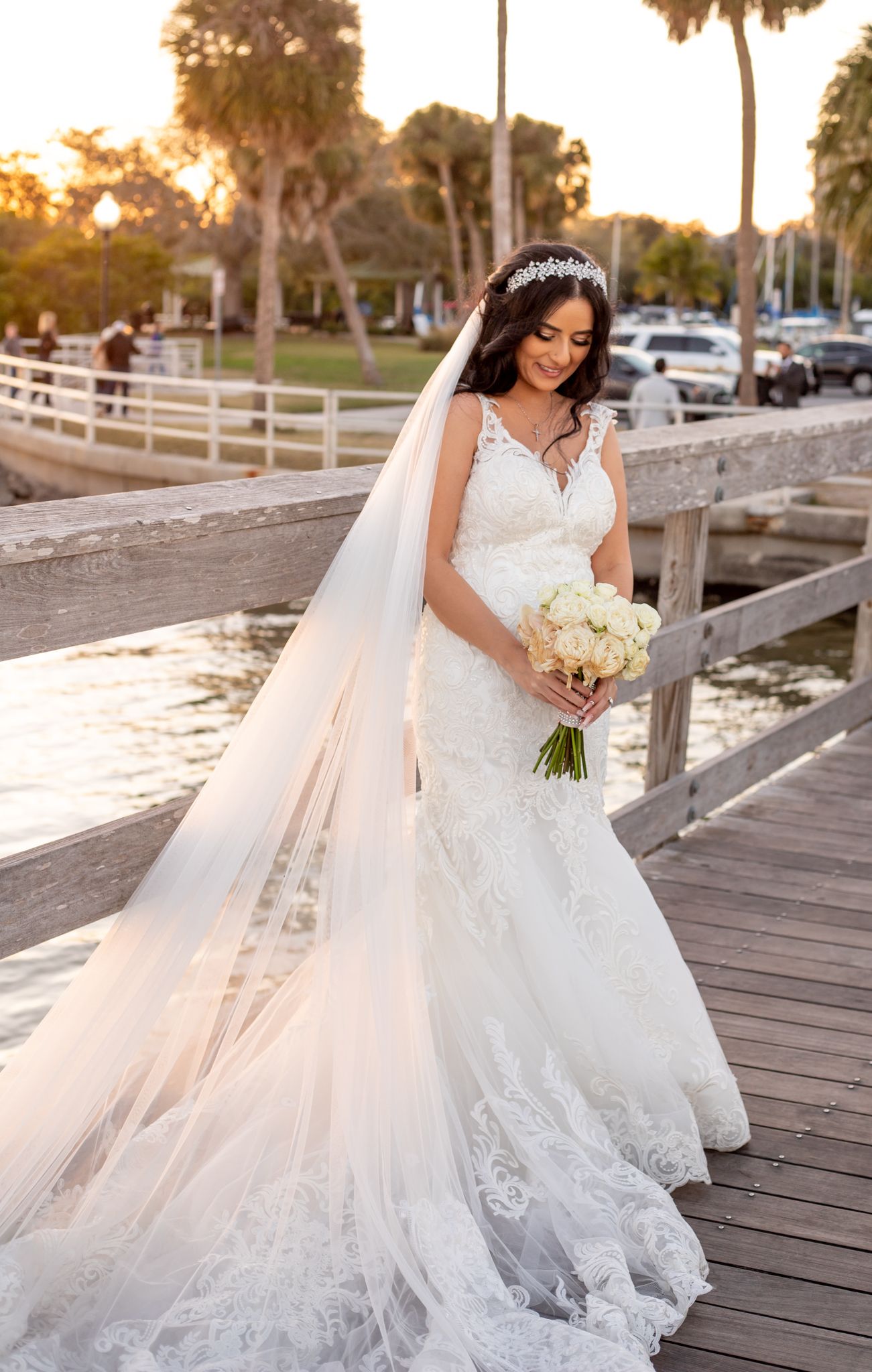 Elegant bride in lace gown and flowing veil poses on wooden boardwalk during golden hour.