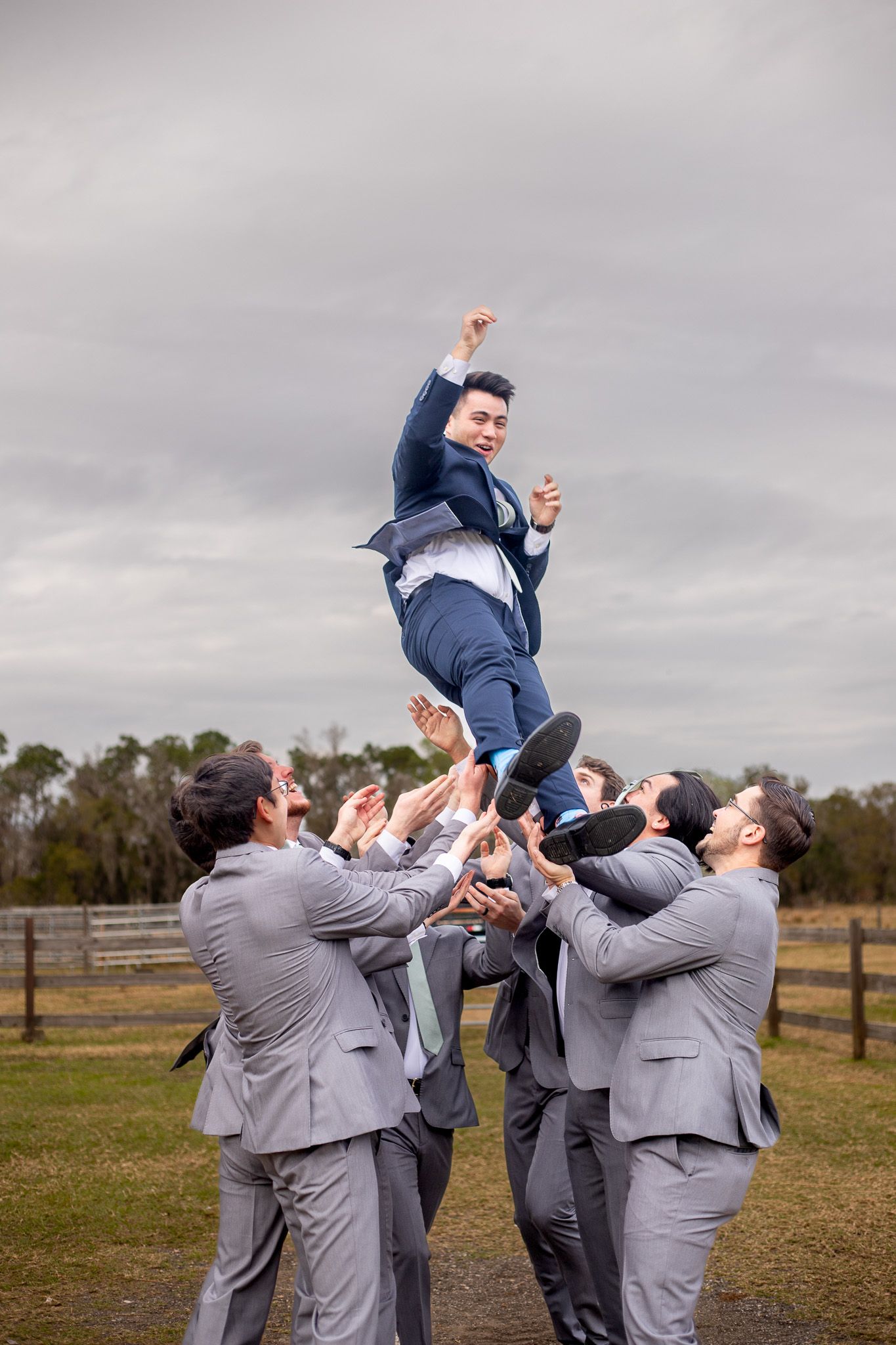 Groomsmen in grey suits playfully toss someone in the air during outdoor wedding photo.