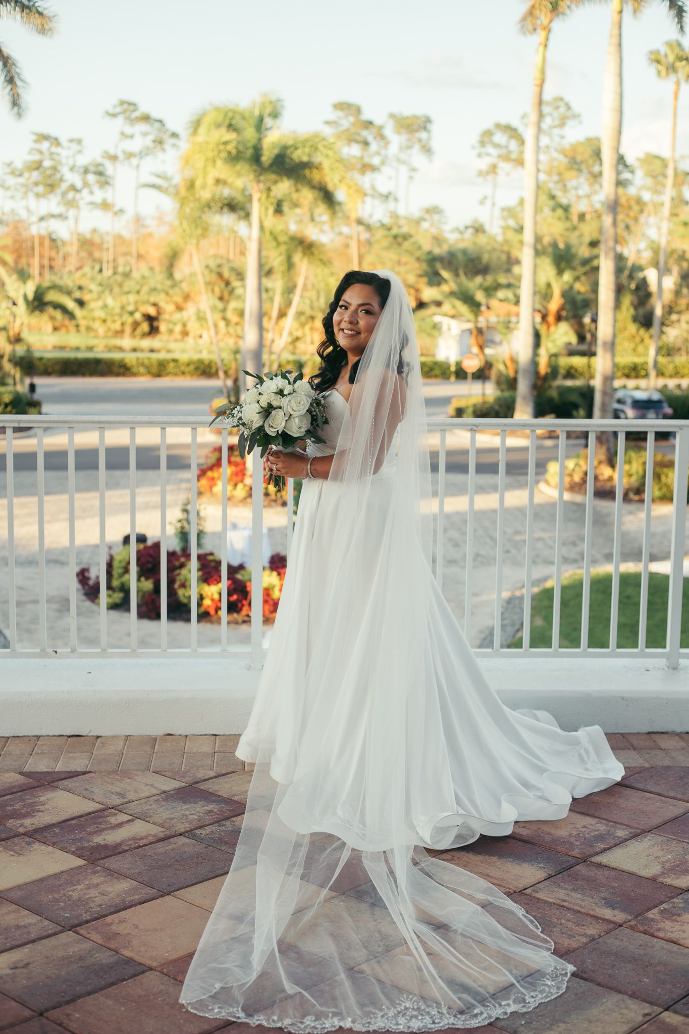 Bride in flowing gown poses on terrace with palm trees during golden hour.