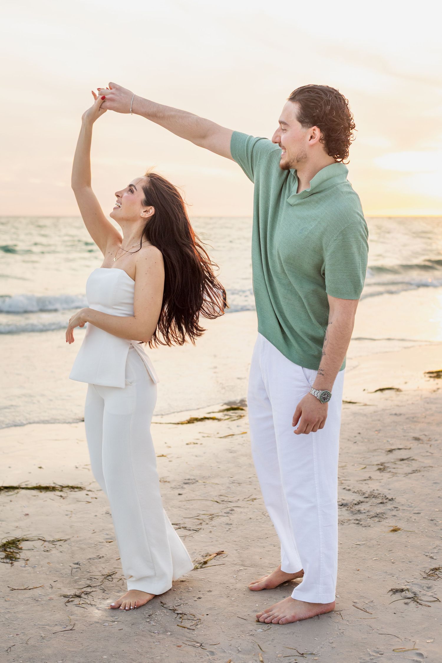 Couple dancing barefoot on a beach at sunset, man spinning woman in white outfit, ocean waves in background.