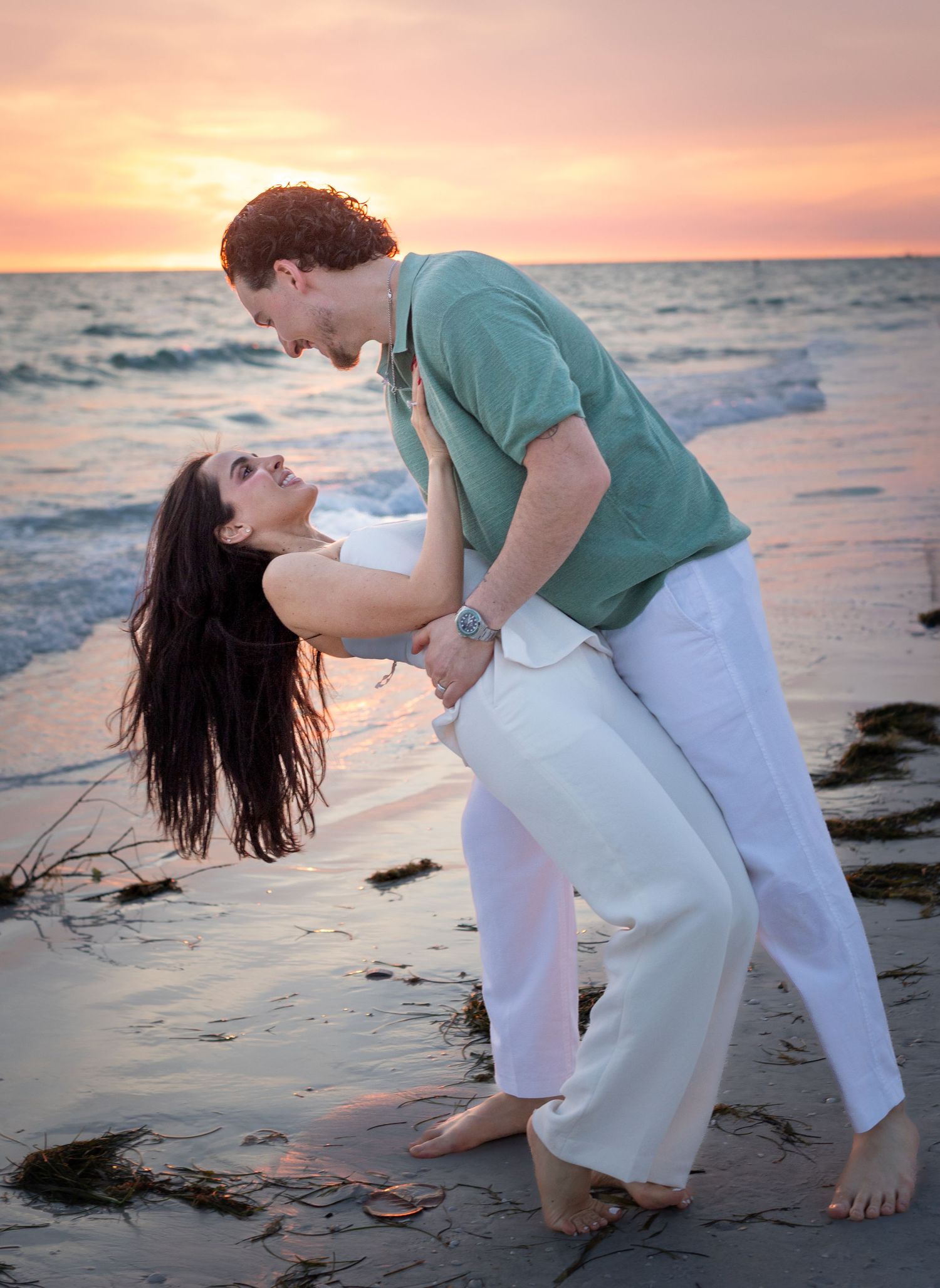 Couple dancing romantically on a beach at sunset, woman leaning back in mans arms, both wearing white pants.
