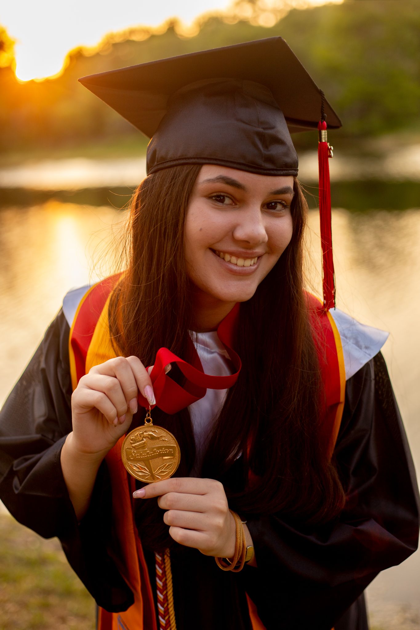 Graduate in academic regalia shows off gold medal during sunset photoshoot.