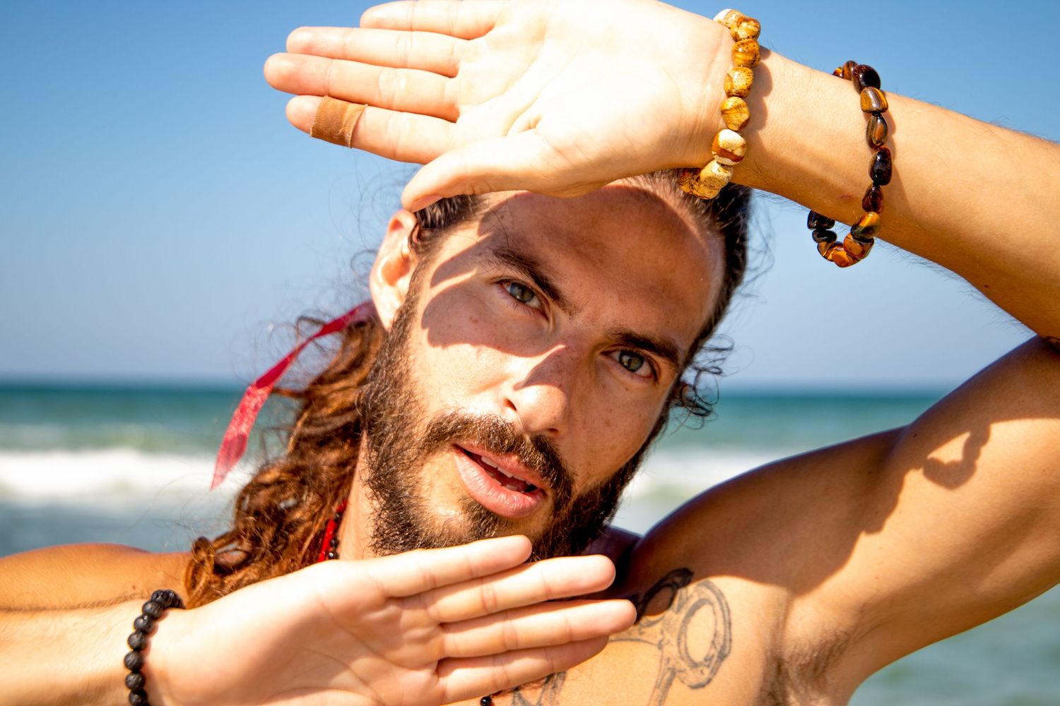 Person with beaded bracelets poses on beach with hands framing face.