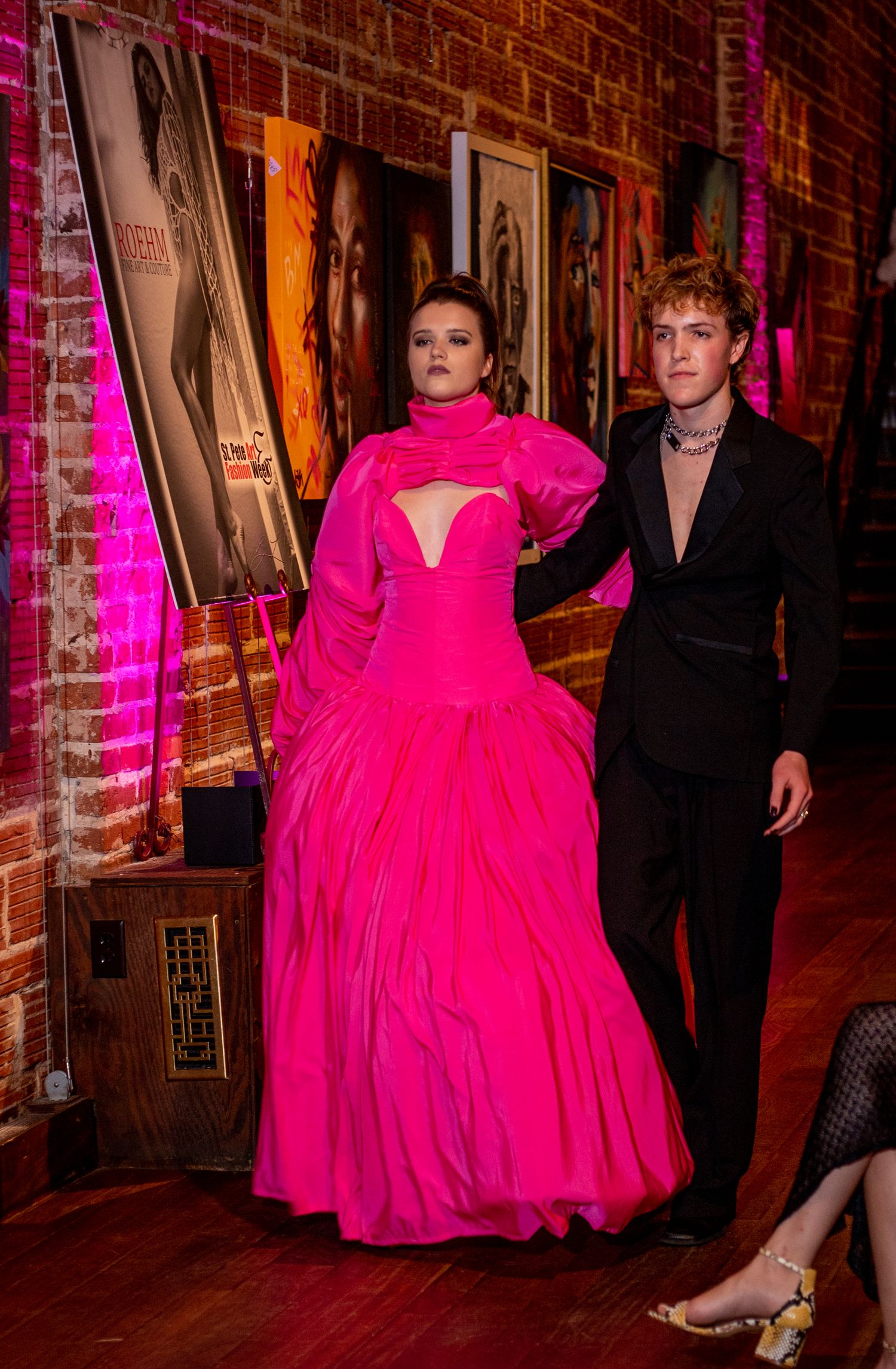 A fashion show duo posing in front of artwork, featuring a pink ballgown and black suit.