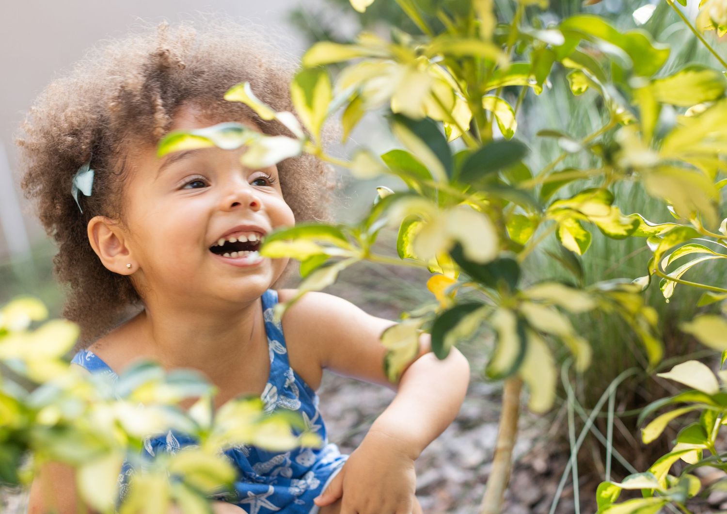 Child smiles brightly while playing among sunlit green foliage.