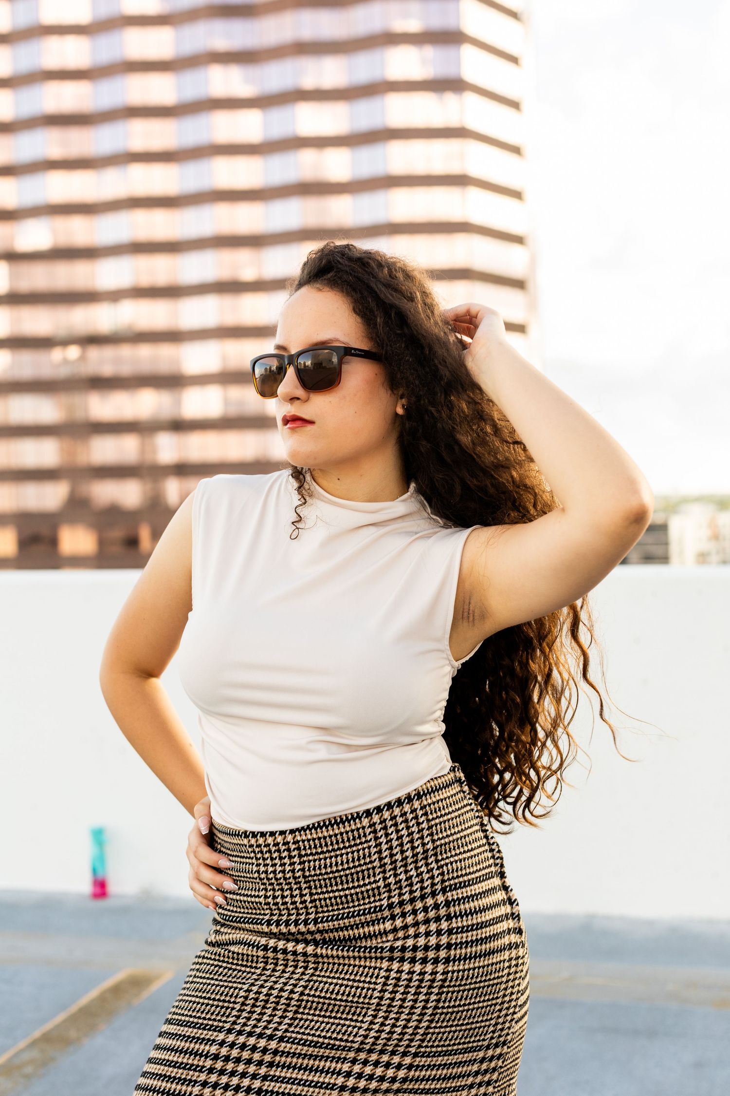 Fashionable woman in a white sleeveless top and plaid skirt poses confidently against an urban backdrop.