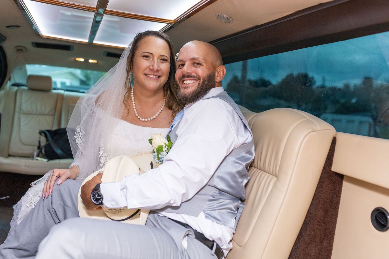 Bride and groom smiling together in a wedding limousine, bride in white gown and veil, groom in gray suit.