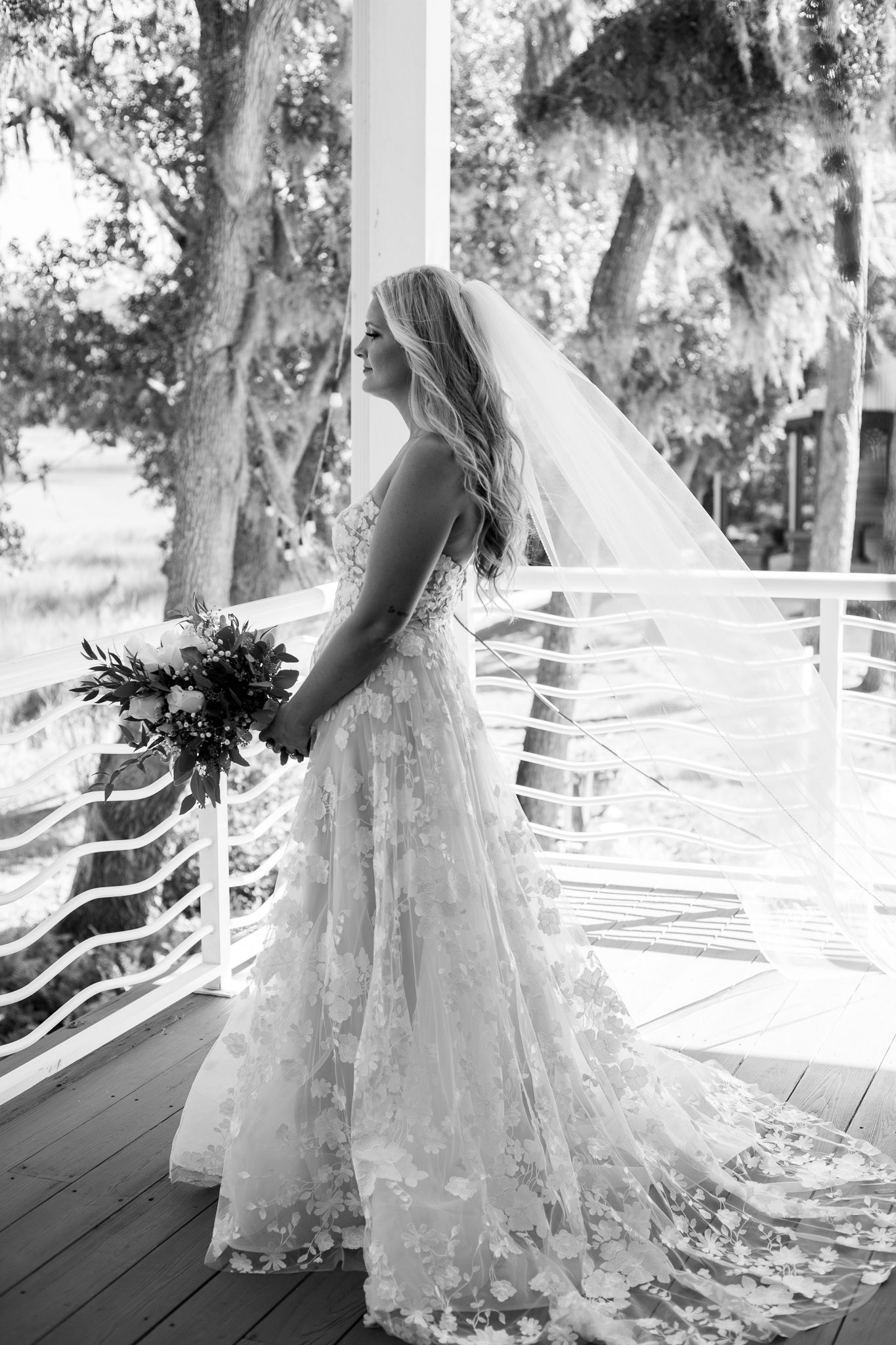 Bride in floral lace gown and veil holds bouquet on porch, black and white wedding portrait.