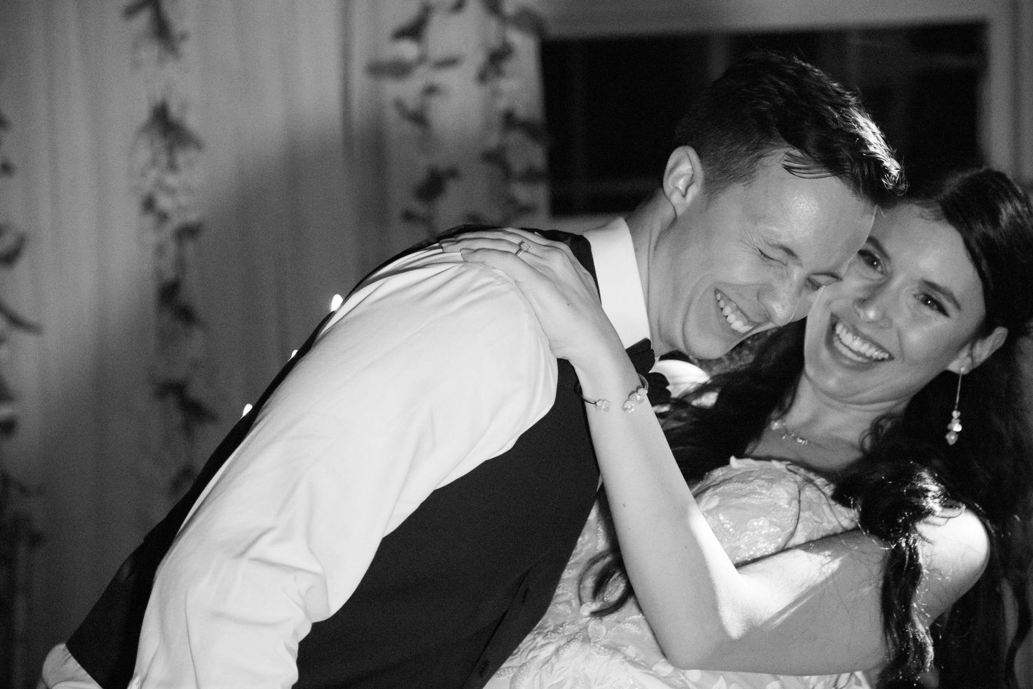 Black and white photo of a laughing bride and groom sharing a joyful embrace during their wedding reception dance.