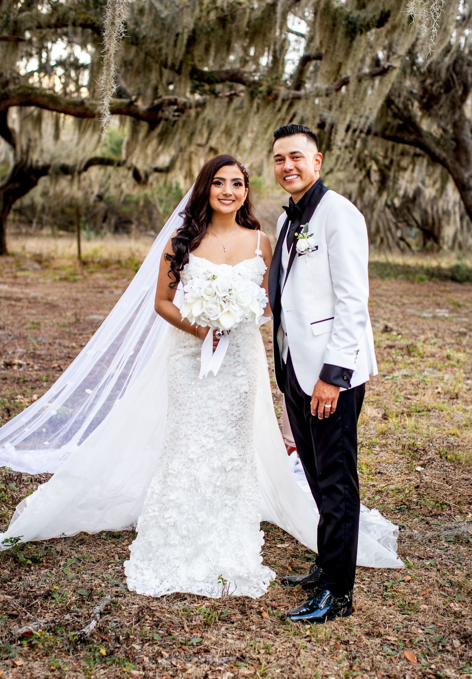 Bride in white lace gown with veil and groom in white tuxedo jacket pose outdoors under moss-draped oak trees.