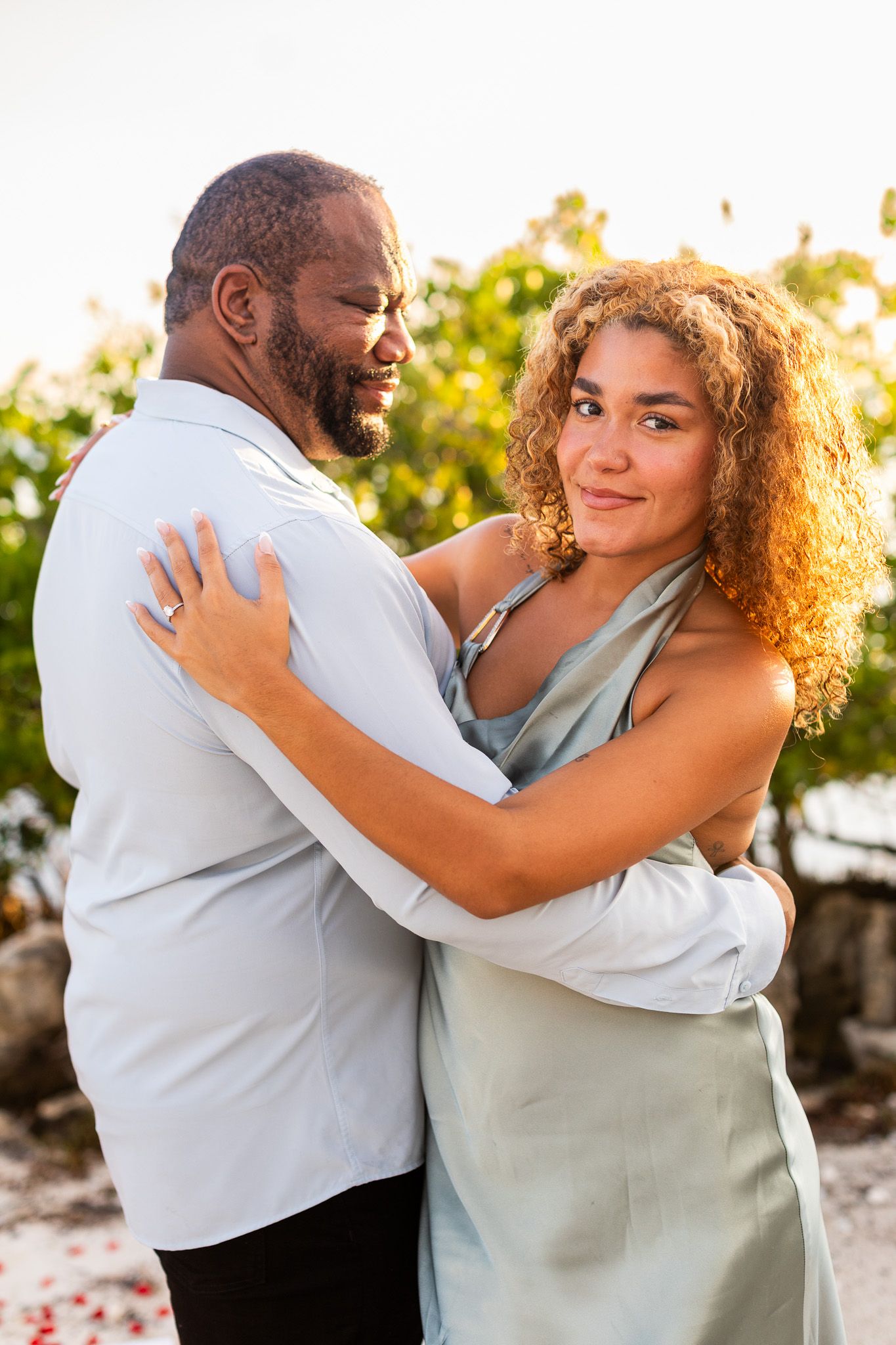 Couple embracing outdoors at sunset, woman in sage green dress with curly hair smiling, man in white shirt, tropical background.