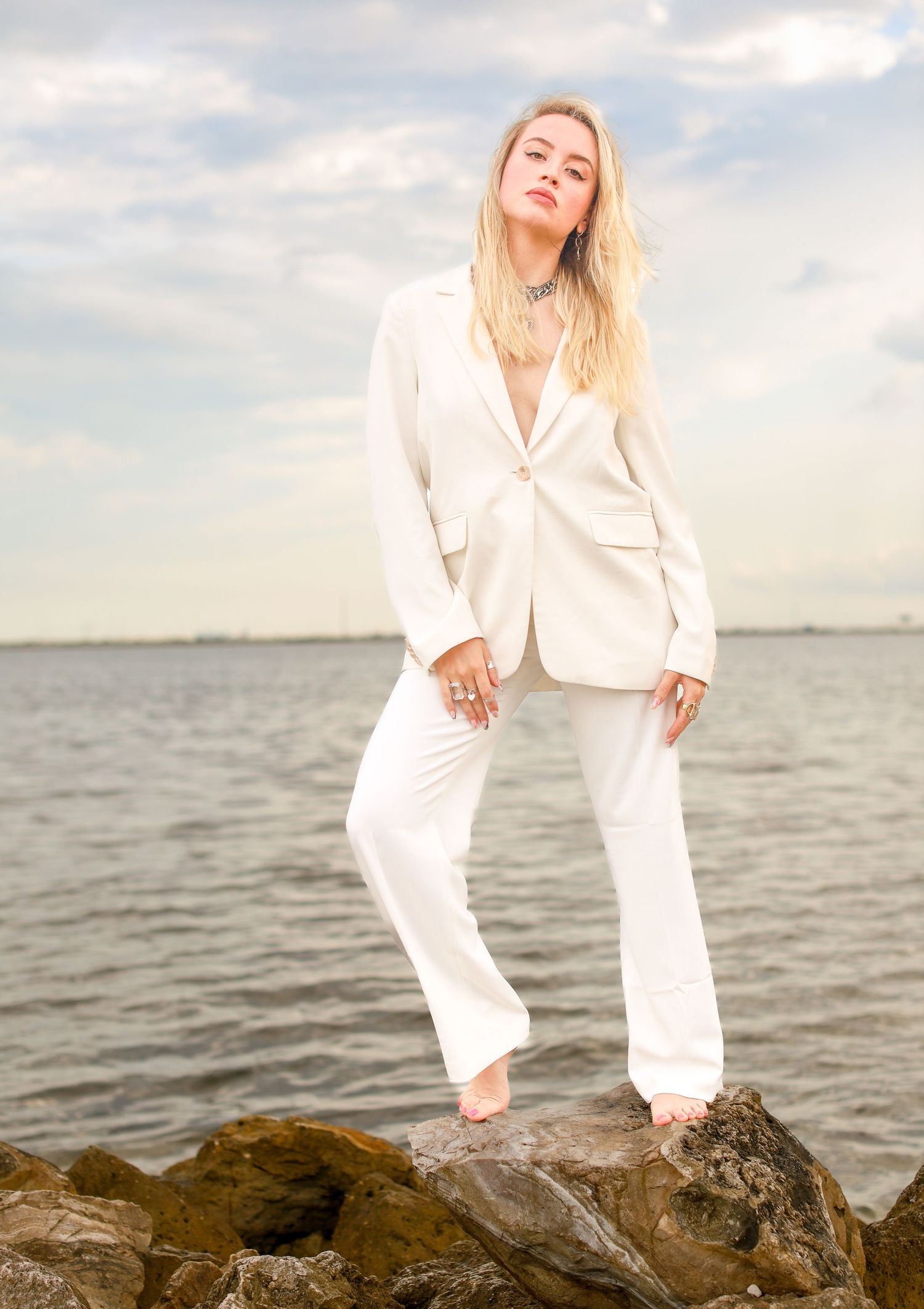 Figure in crisp white suit poses confidently on coastal rocks at sunset.