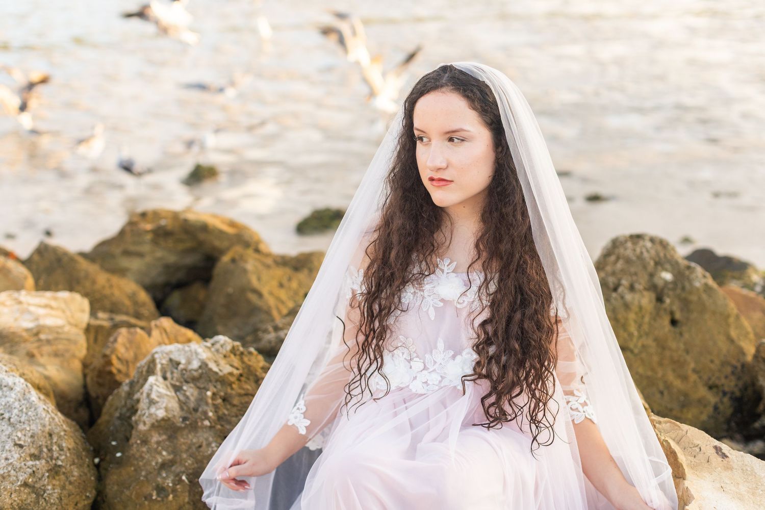 Seaside portrait featuring long curls and white lace dress against rocky coastal backdrop.
