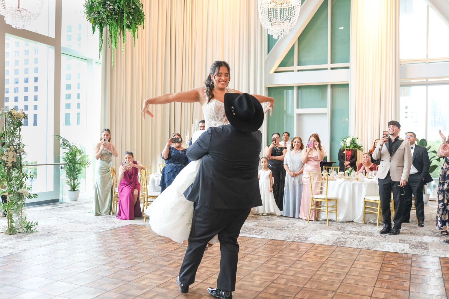 Bride and groom share first dance as groom lifts bride at elegant wedding reception with guests watching.