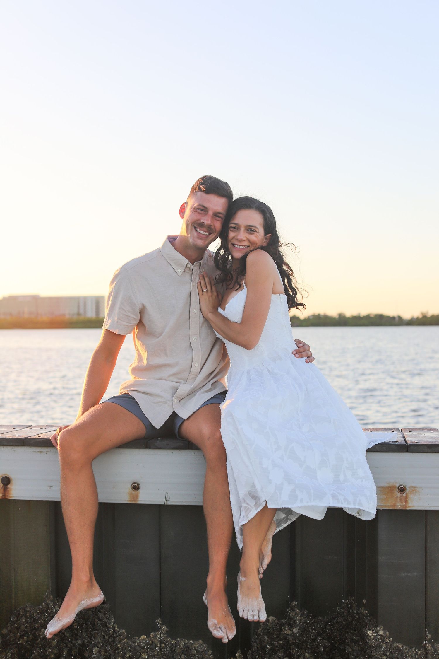 Happy couple sitting on a waterfront dock at sunset, woman in white dress and man in casual shirt, smiling together.