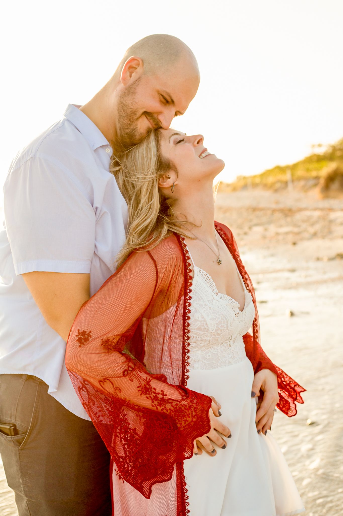 A romantic couple embraces on the beach at sunset wearing white and red outfits.