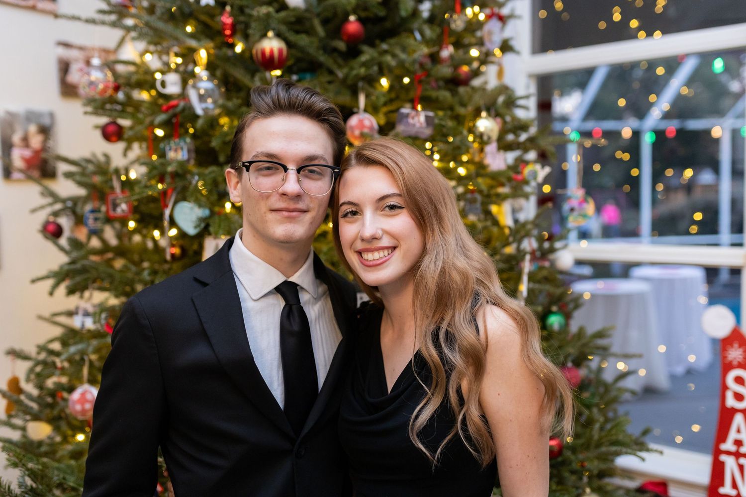 Couple poses in front of decorated Christmas tree with ornaments and lights in elegant holiday setting.