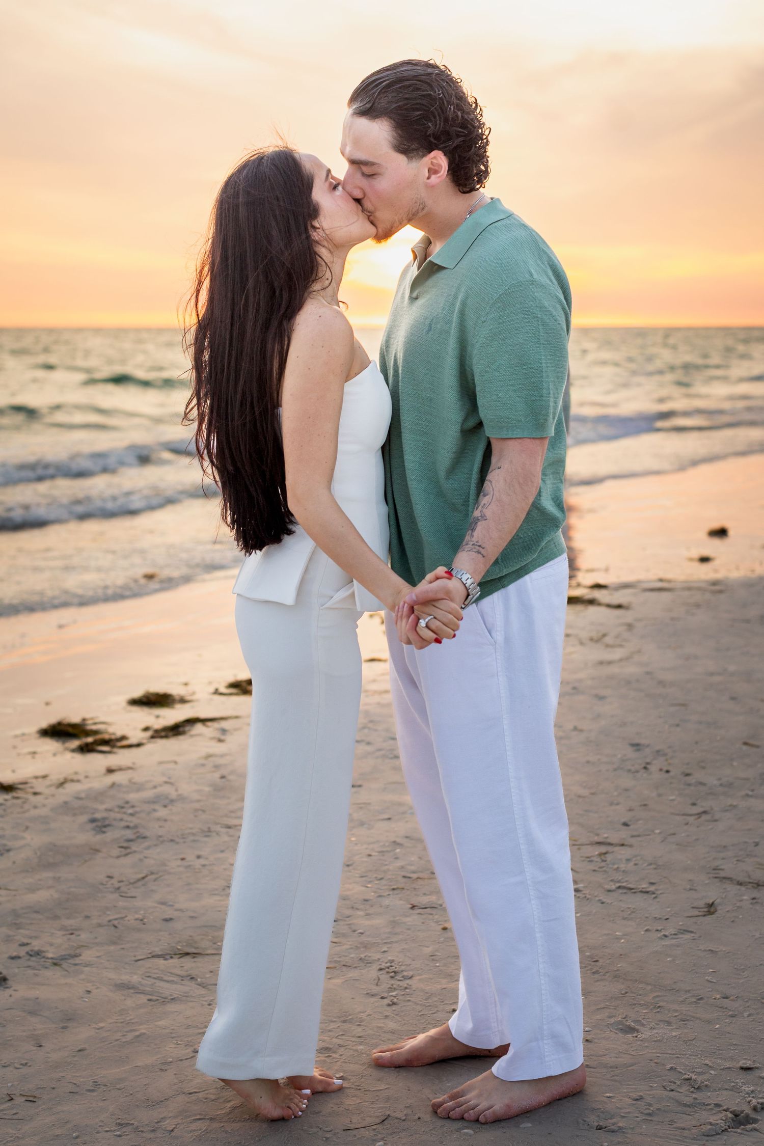 Couple kissing on beach at sunset, woman in white outfit and man in green shirt holding hands by the ocean.