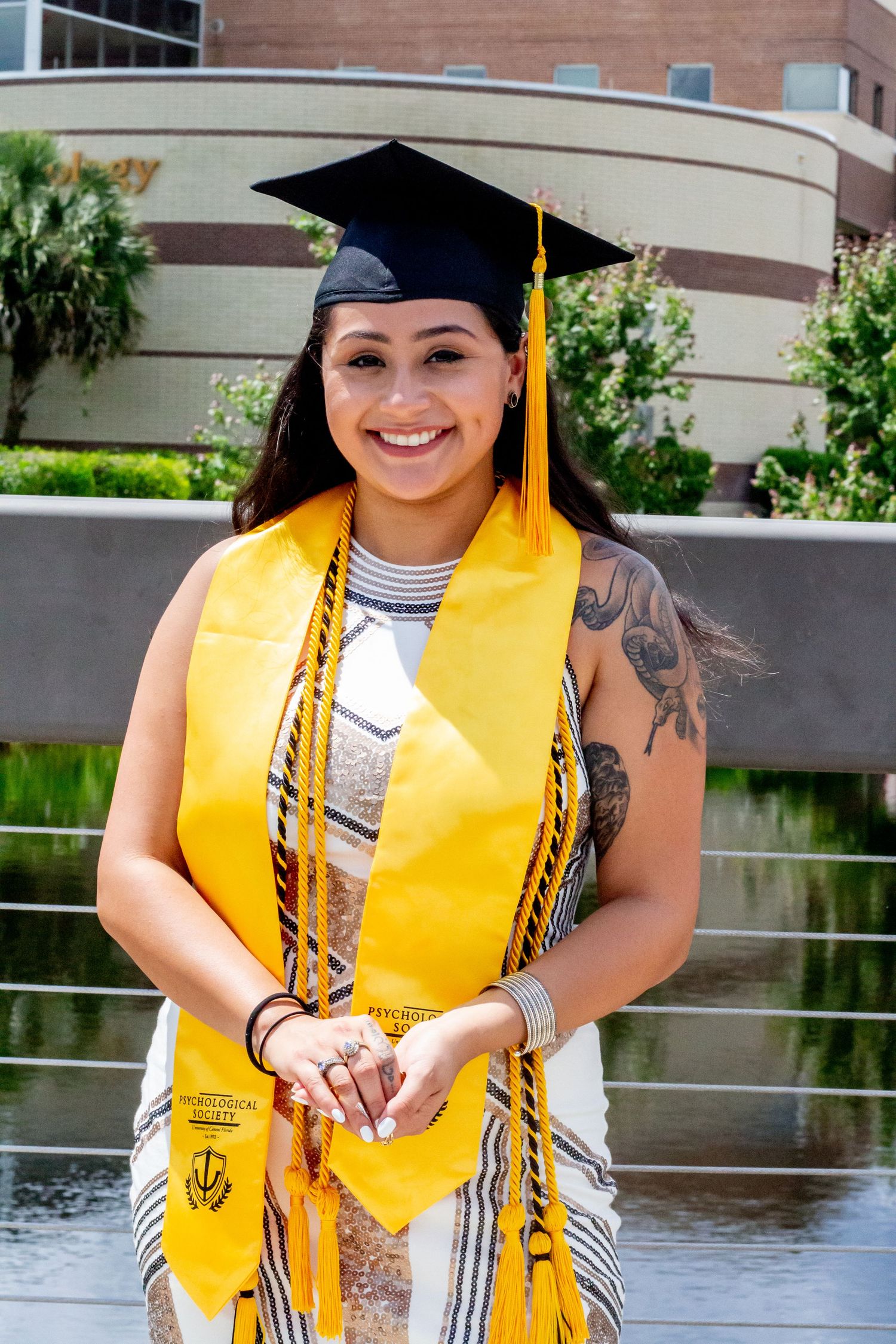 Graduate in traditional cap and gown with gold stole poses by reflecting pool.