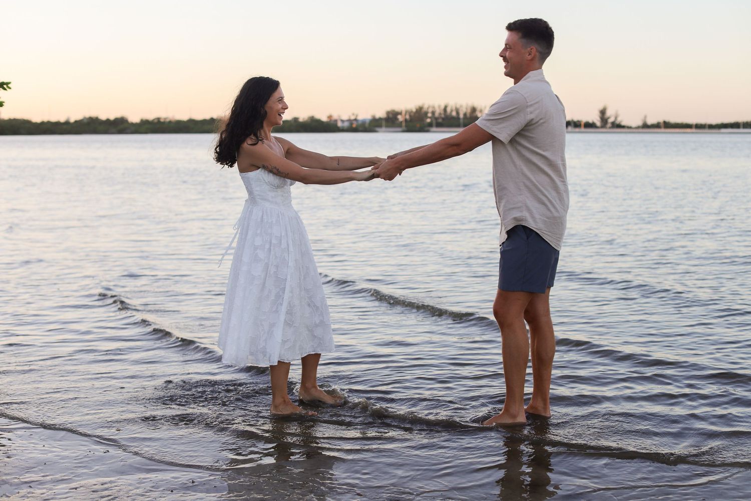 Couple holding hands standing barefoot in shallow water at sunset near a serene lake shore.