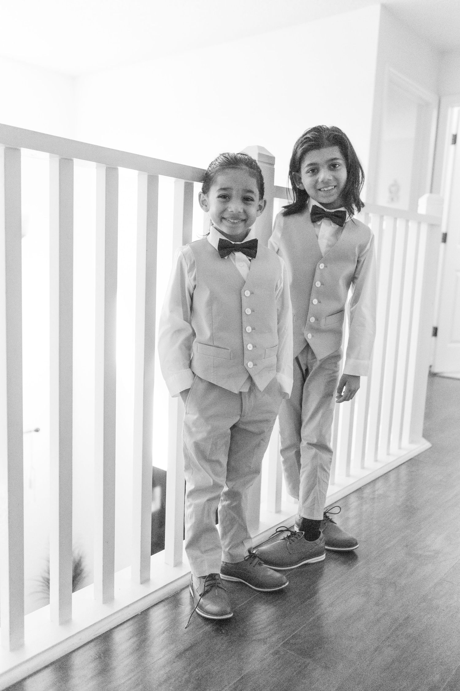 Two young boys in matching bow ties and vests pose smiling by white stair railing in black and white photo.