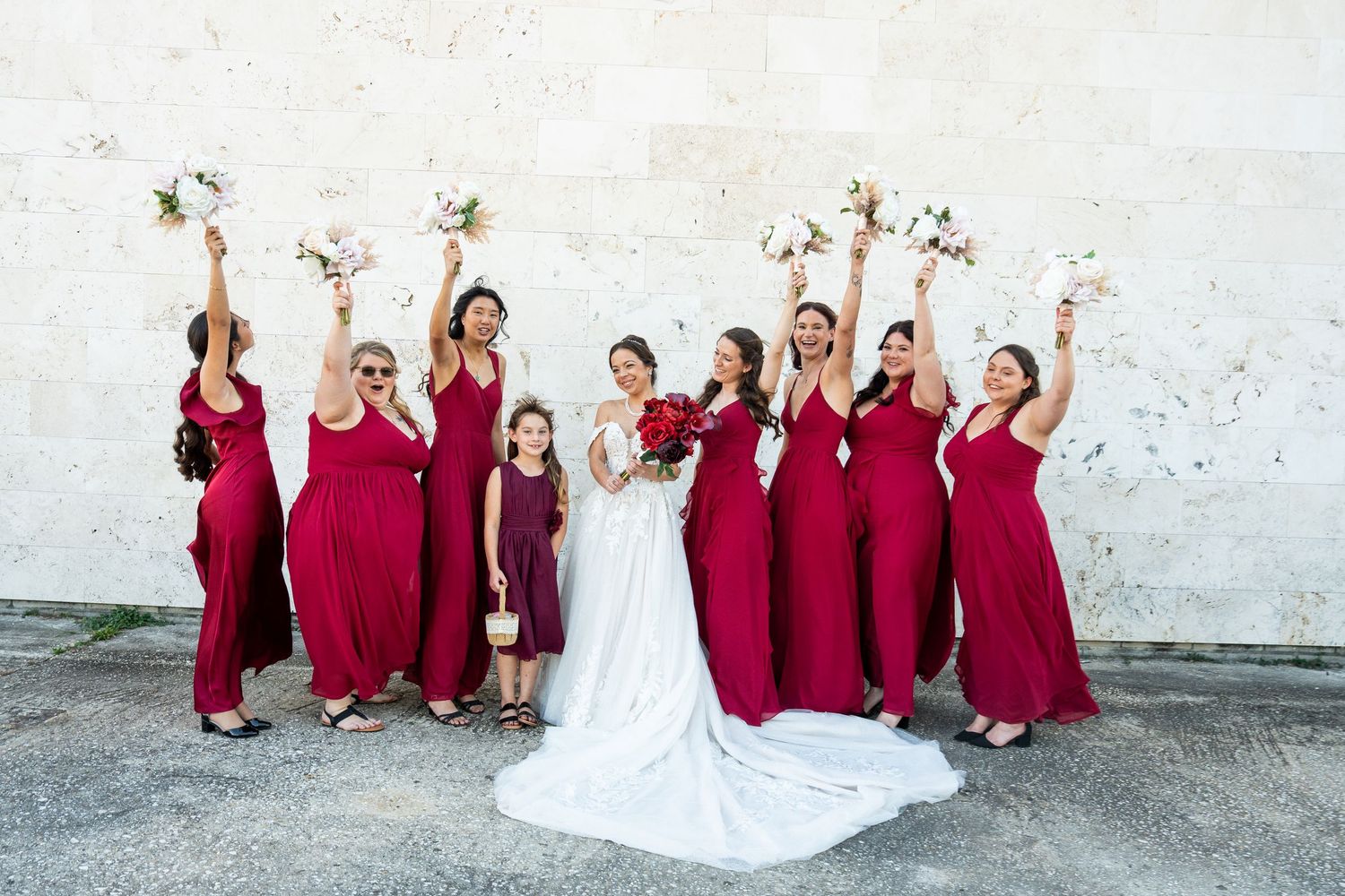 Bride in white gown with bridesmaids in red dresses raising bouquets outdoors against a white wall.