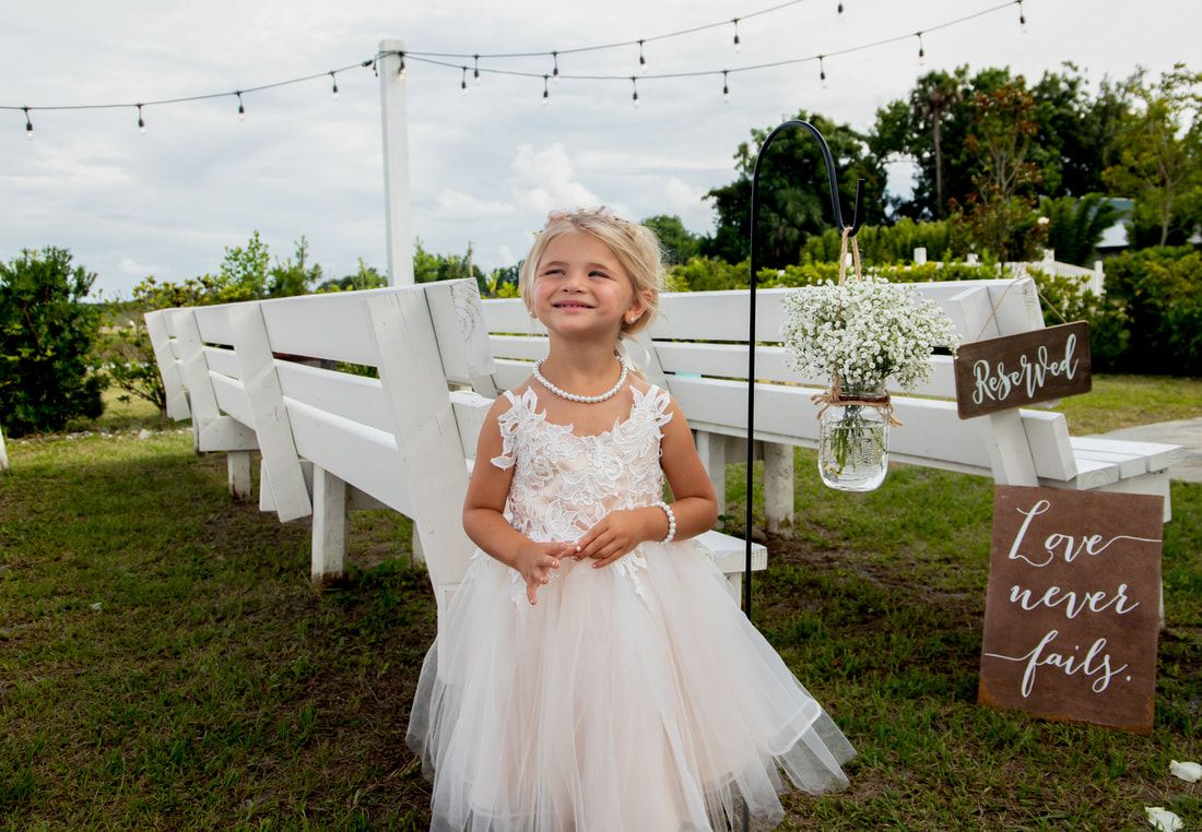 Young flower girl in white lace tulle dress stands at outdoor wedding ceremony with white benches and love never fails sign.