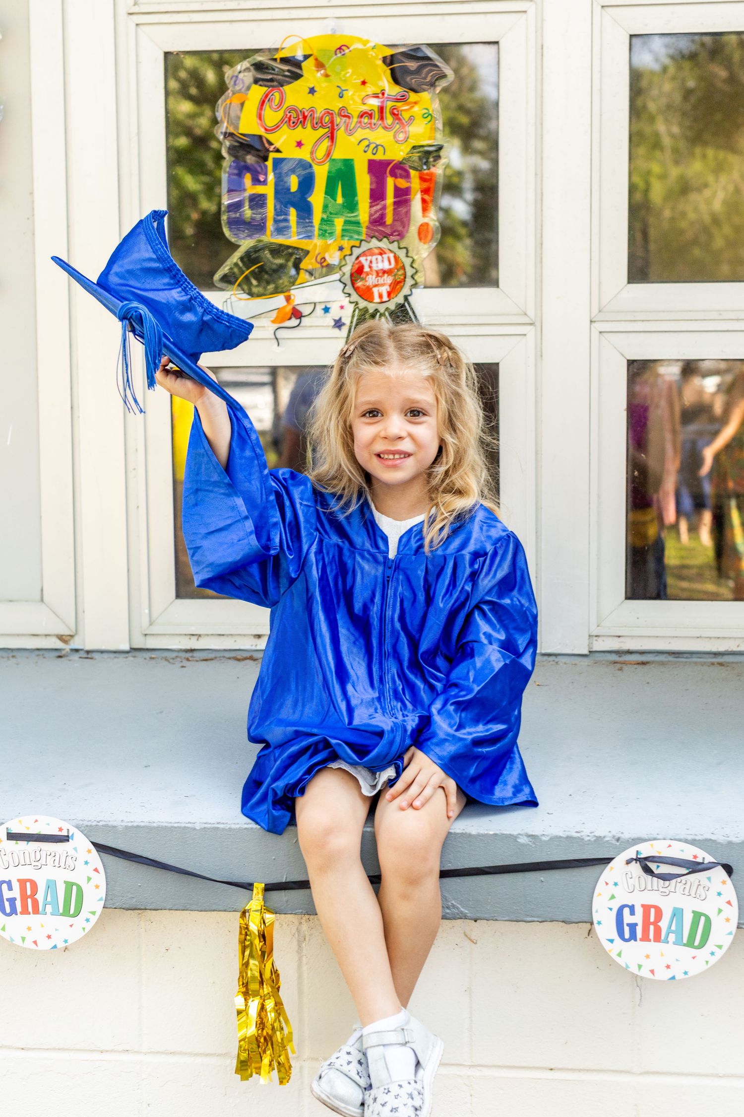 Graduate in blue cap and gown sits on steps with graduation decorations and balloons.