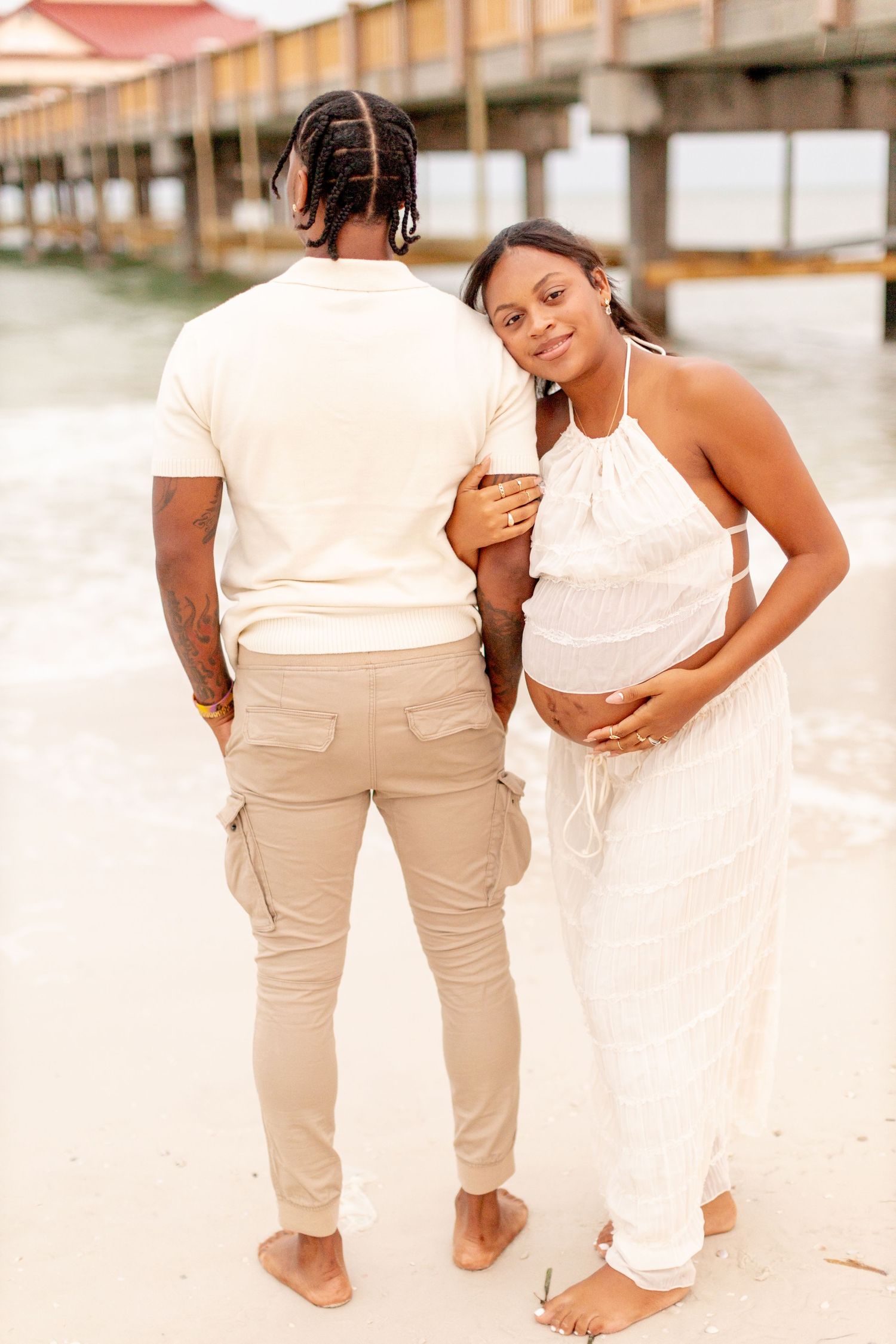 A couple in light summer attire poses together on a sandy beach near a pier.
