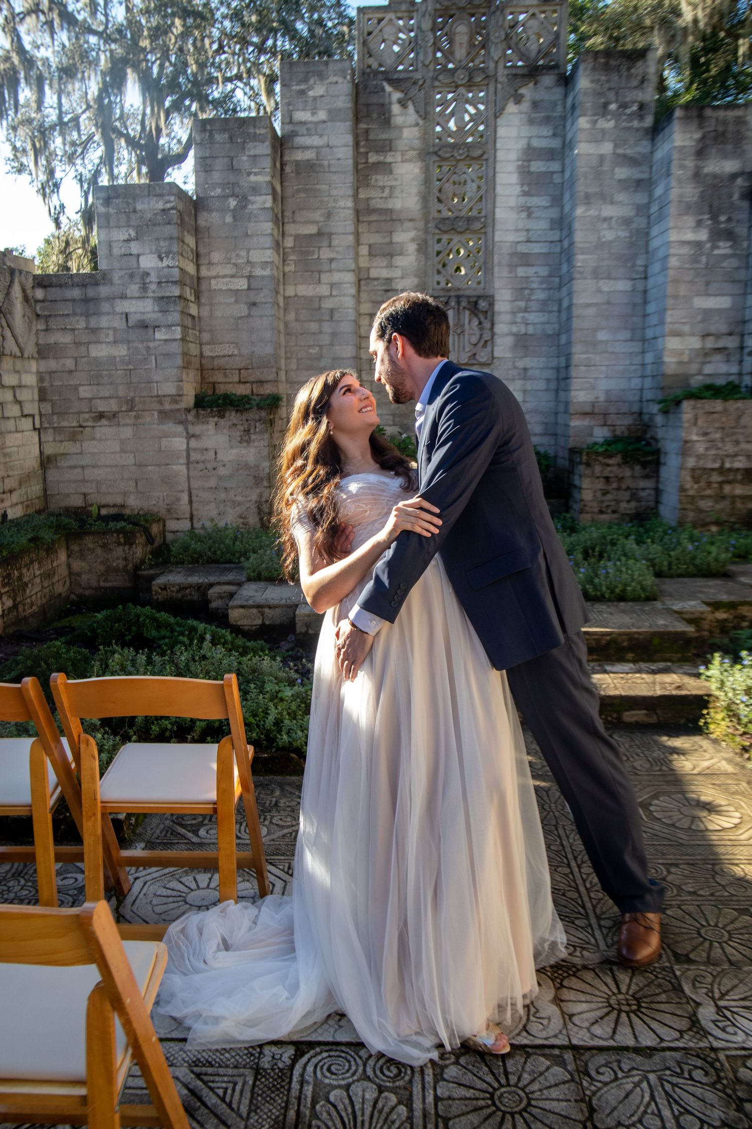 A bride and groom share a romantic dance in an outdoor garden ceremony venue with stone walls and mosaic tile flooring.
