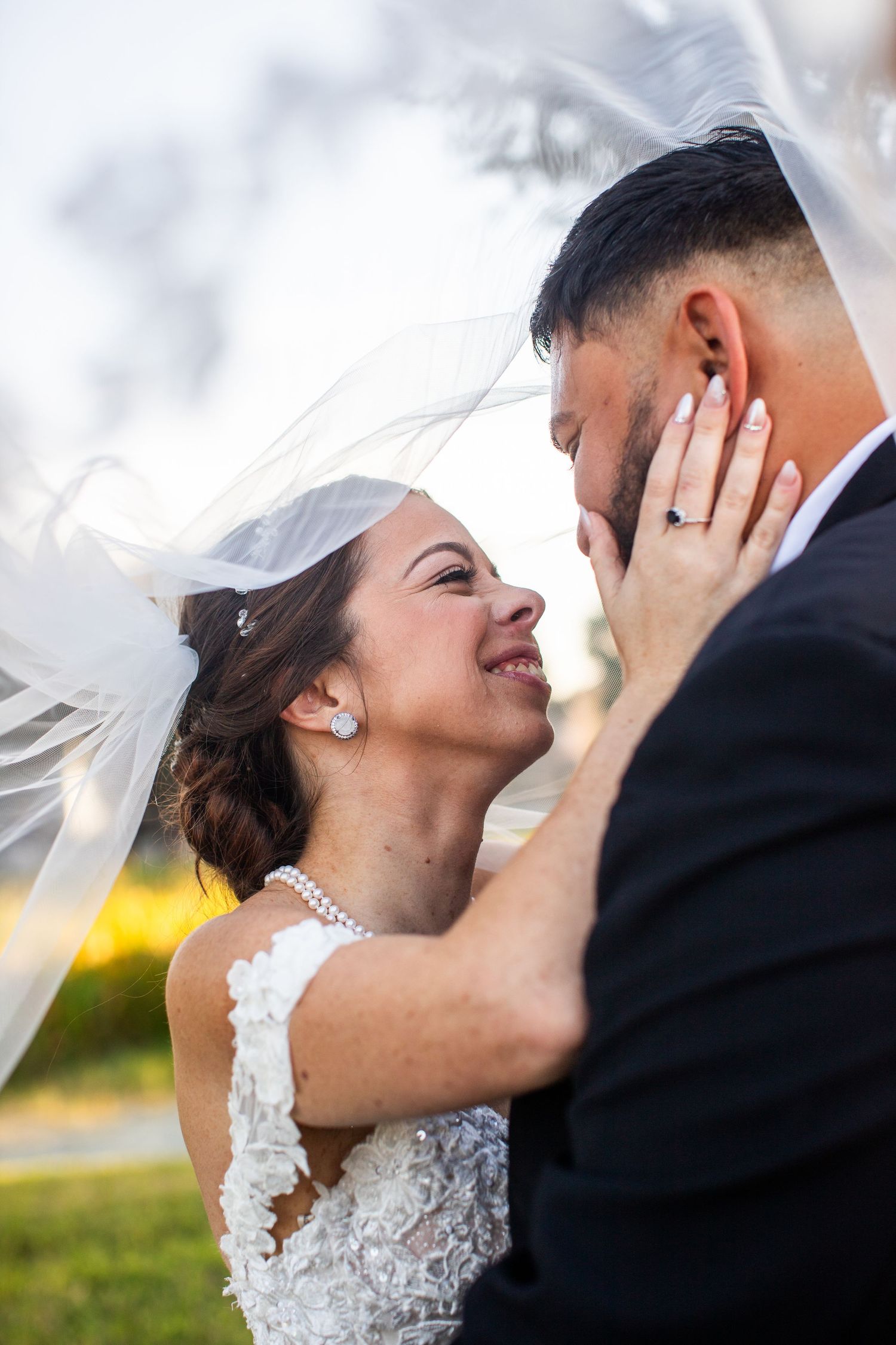 Bride and groom share a romantic moment under a flowing veil, with the bride smiling and touching the groom's face.