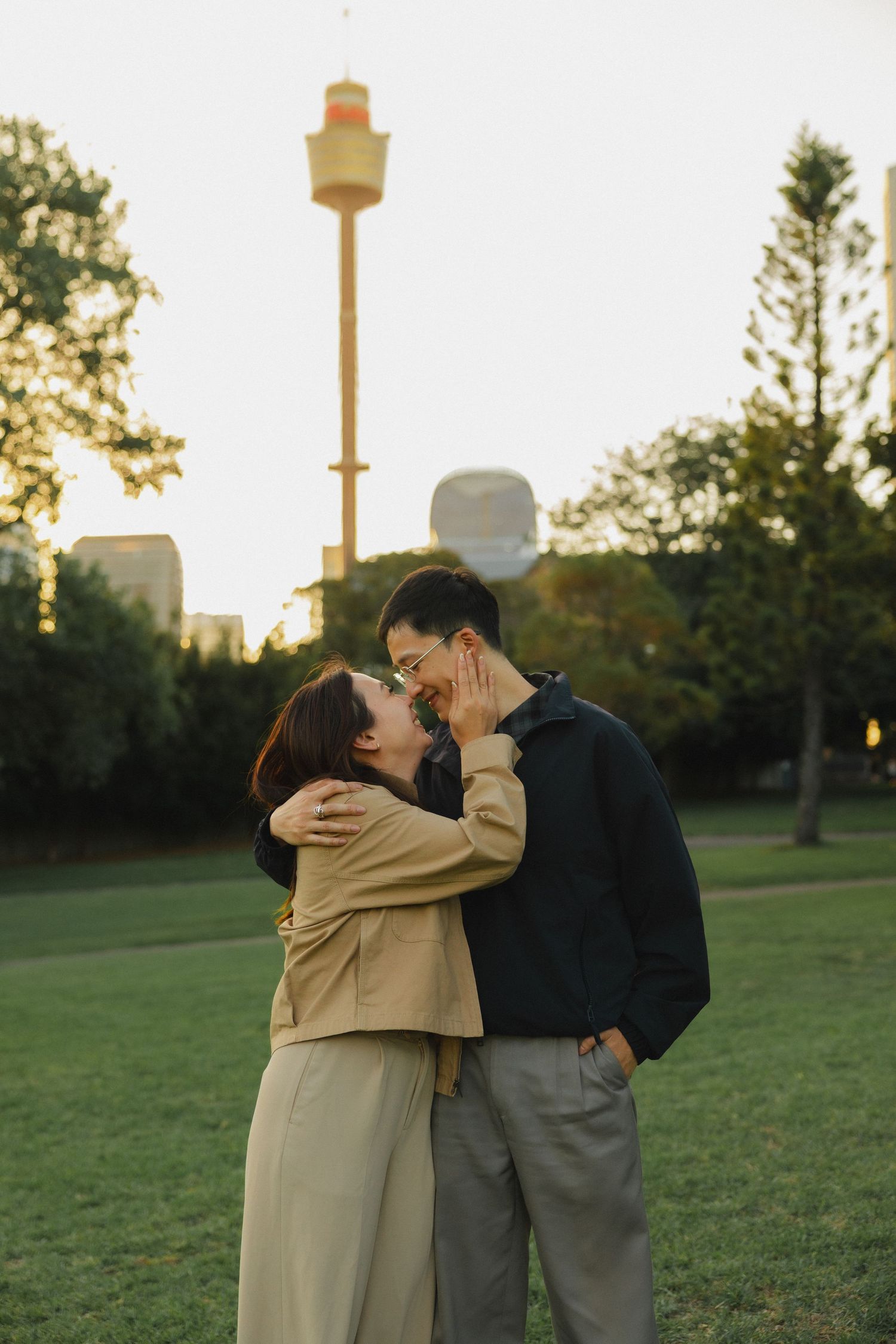 A romantic couple embraces in a park at sunset with Sydney Tower Eye in the background.