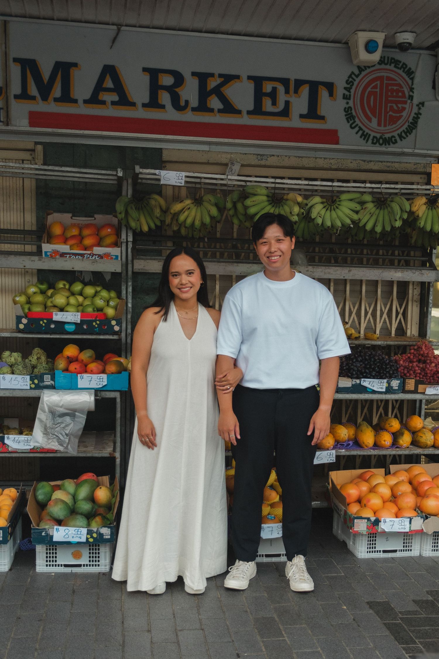 A couple standing in front of a market storefront with fresh fruit and produce displays.