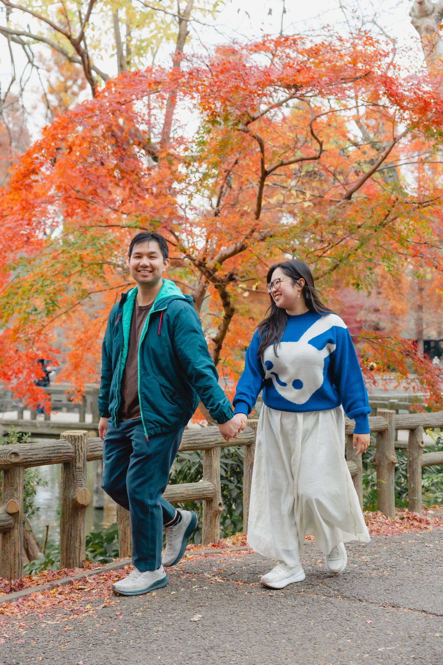 A couple poses playfully in a park with vibrant red autumn foliage and a wooden fence in the background.