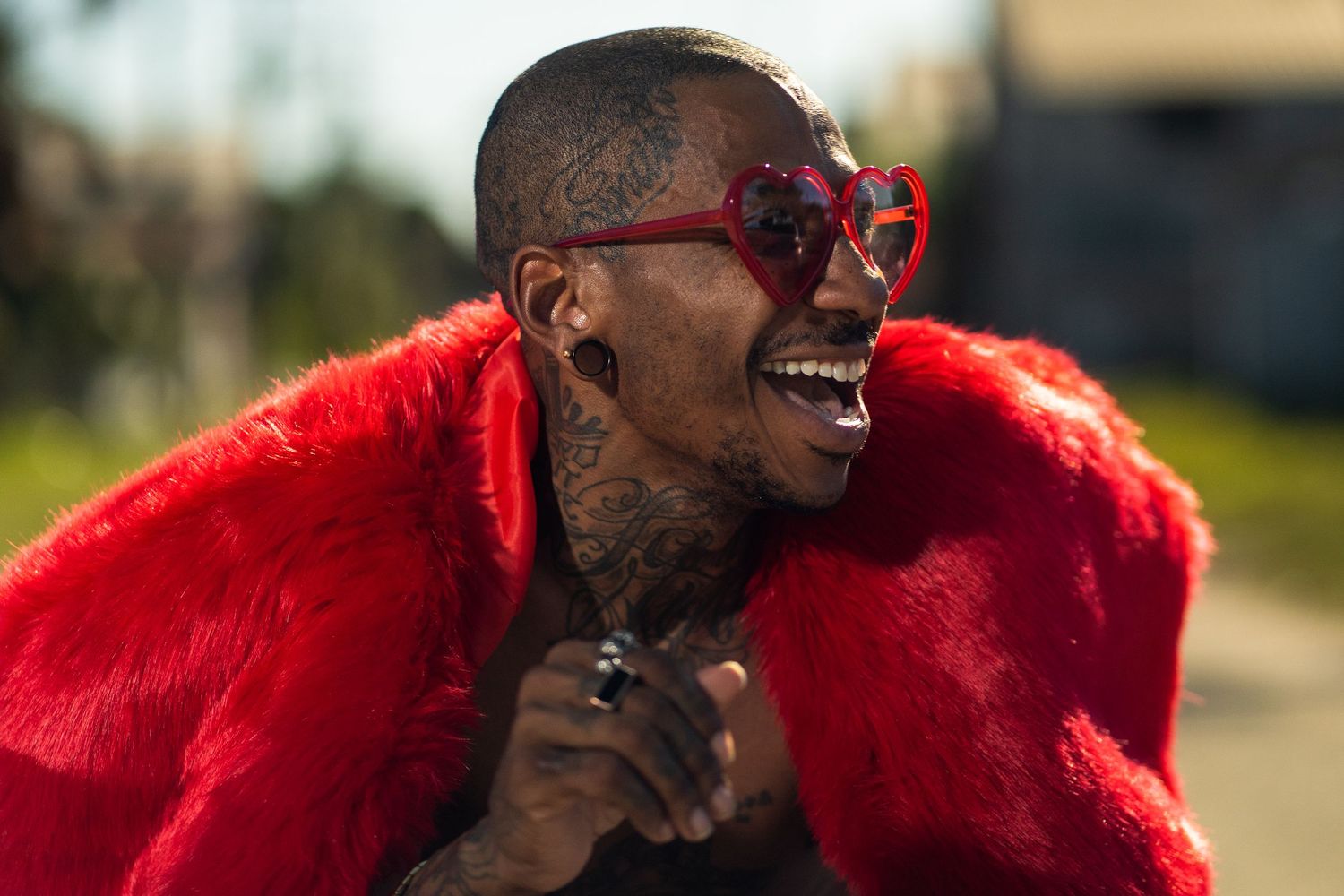Man laughing joyfully while wearing red fur coat and oversized sunglasses.