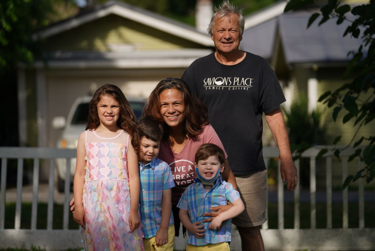 Family portrait outdoors with three children and two adults smiling in front of a house.