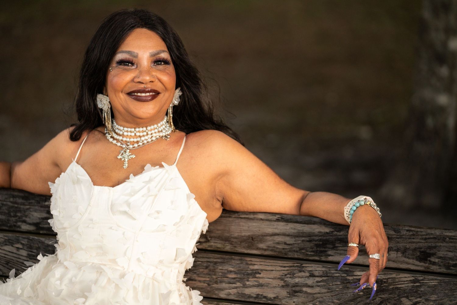 Bride in a lace gown relaxing on a rustic bench with jewelry and smile.