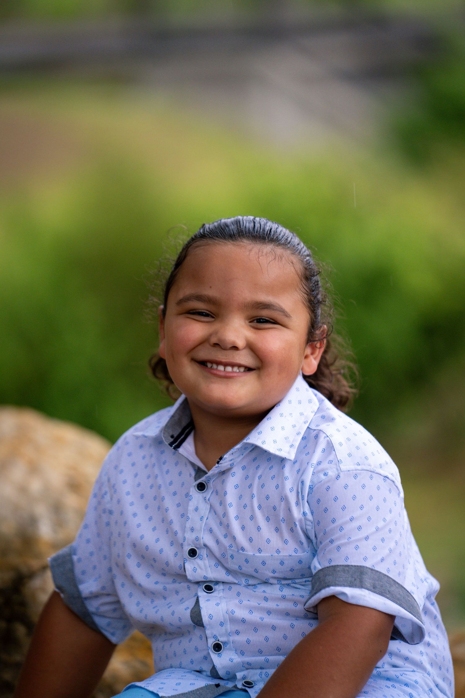 Young boy in a button-down shirt smiling outdoors with blurred greenery.