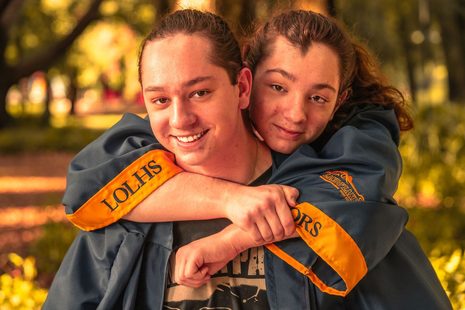 Two graduates in cap and gown hugging and smiling together outdoors.