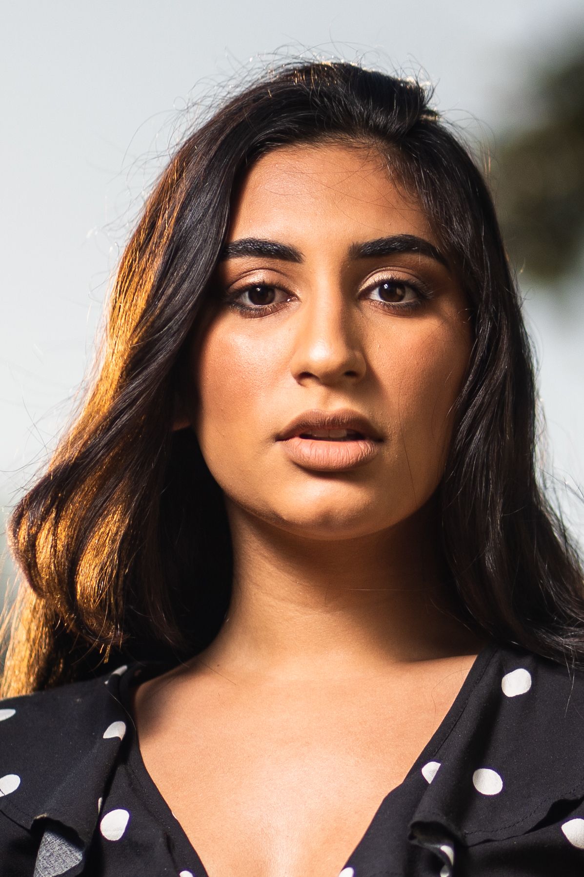 Woman in polka-dot blouse looking directly into the camera with serious expression.