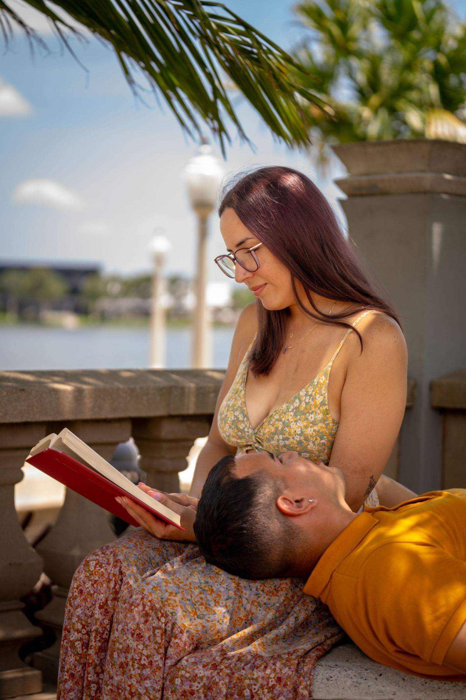 Woman sitting outdoors with a book, enjoying sunshine and shade from palm leaves.