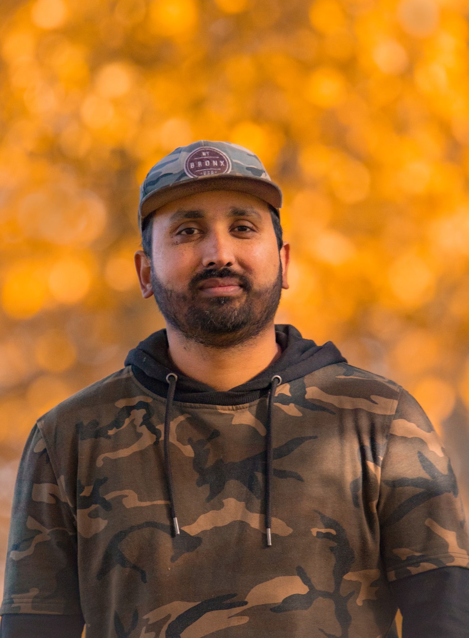 Man with beard wearing camo hoodie and snapback cap posing outdoors against golden bokeh background.