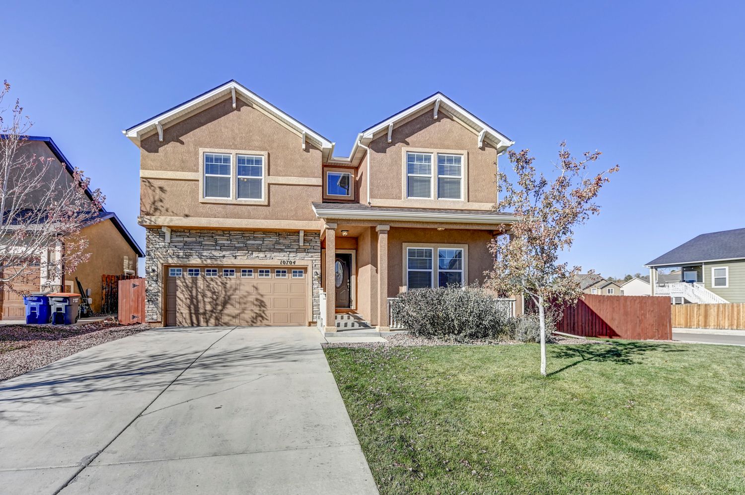 Two-story tan stucco home with attached garage, stone accents, and a well-maintained front lawn under blue skies.