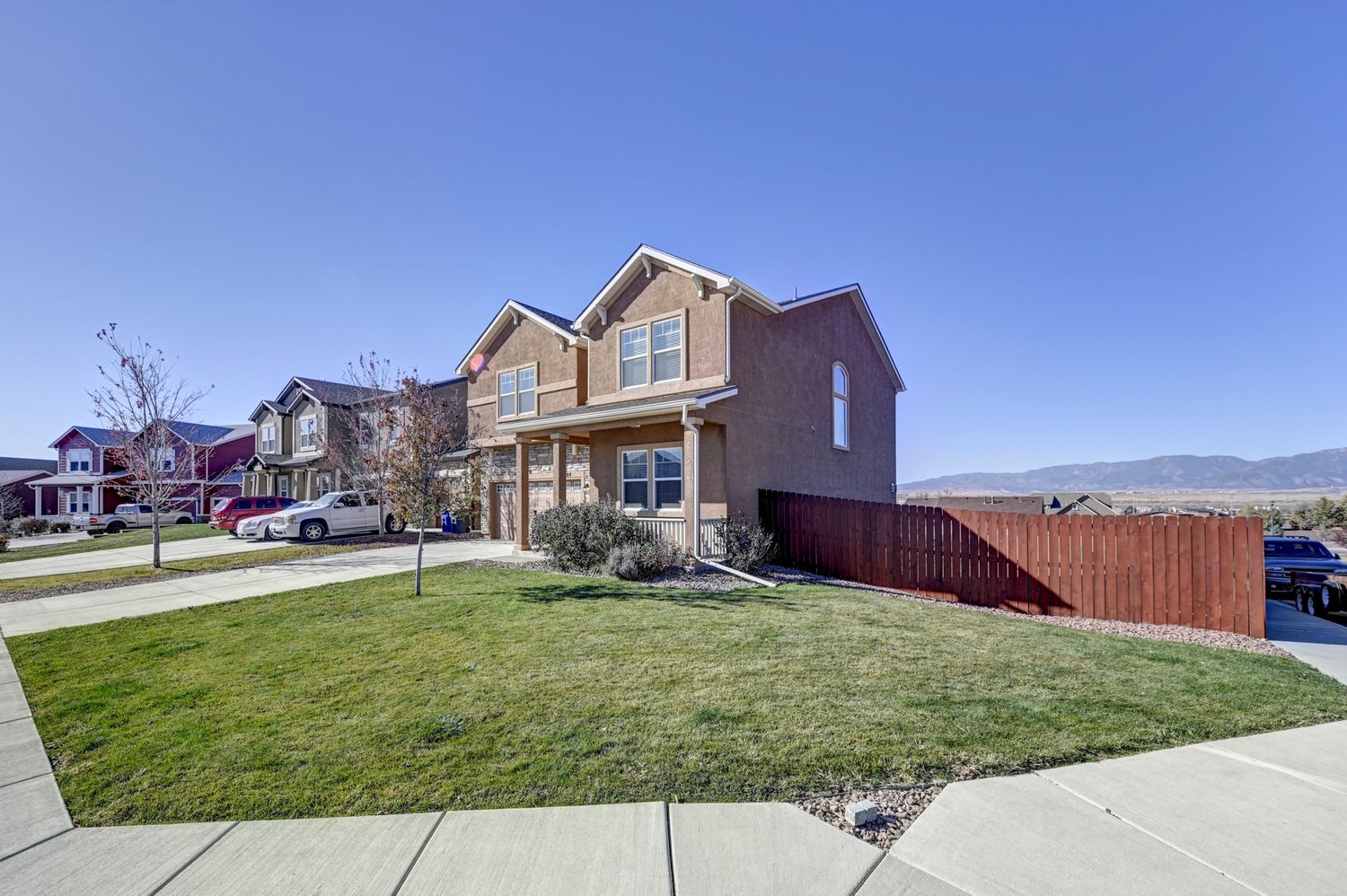 Two-story brick corner home with attached garage, green lawn, wood fence, and mountain views in the background.