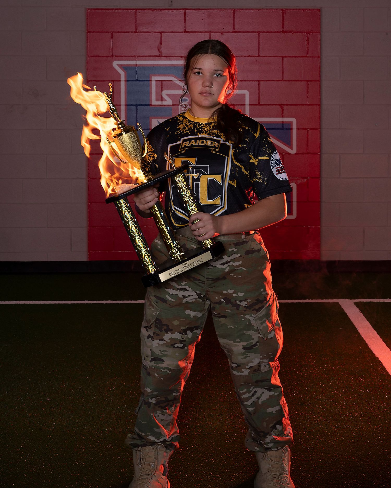 Young athlete in camo pants holds flaming trophy wearing Raiders FC jersey against red brick wall.