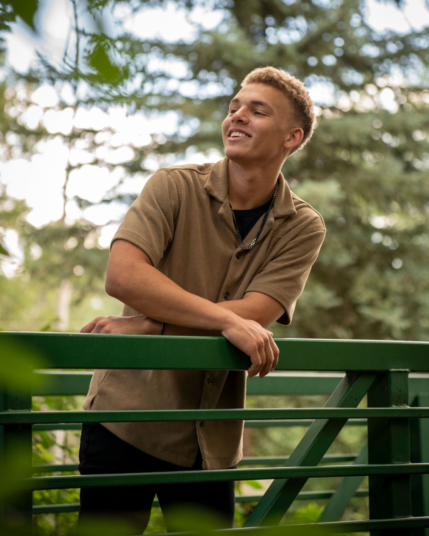 Smiling young man leaning on a green metal fence outdoors surrounded by lush green trees.