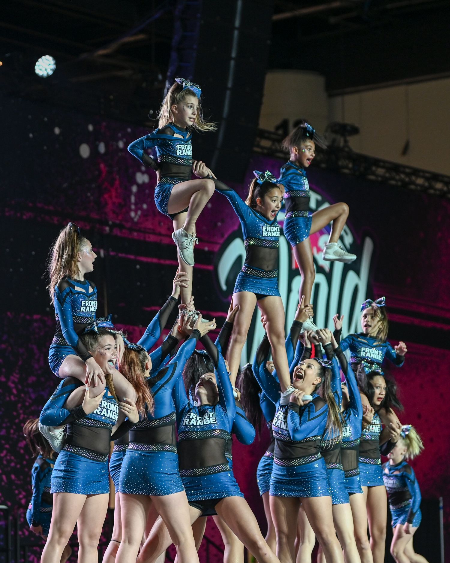Competitive cheerleading squad in blue uniforms performing a multi-level stunt pyramid on stage.