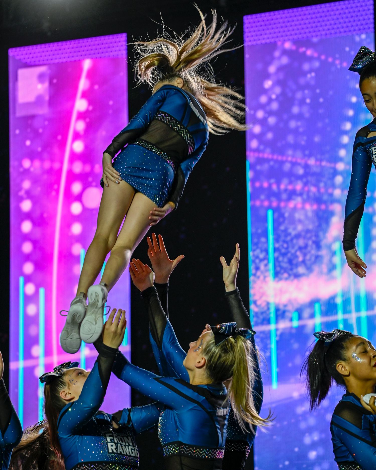 Cheerleader being tossed in air by teammates in blue sequin uniforms under colorful stage lighting.